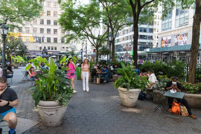 Two women walk through Greeley Square Park amid flowers and trees in New York City.