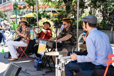 Musicians are seated outdoors in Greeley Square Park playing horns and keyboards.