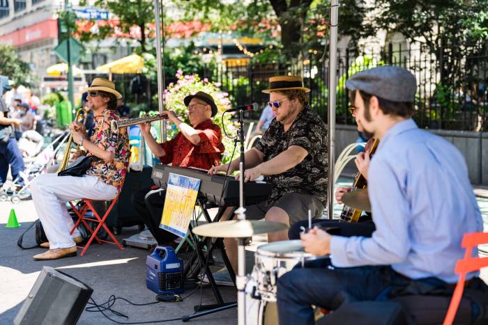 Musicians are seated outdoors in Greeley Square Park playing horns and keyboards.